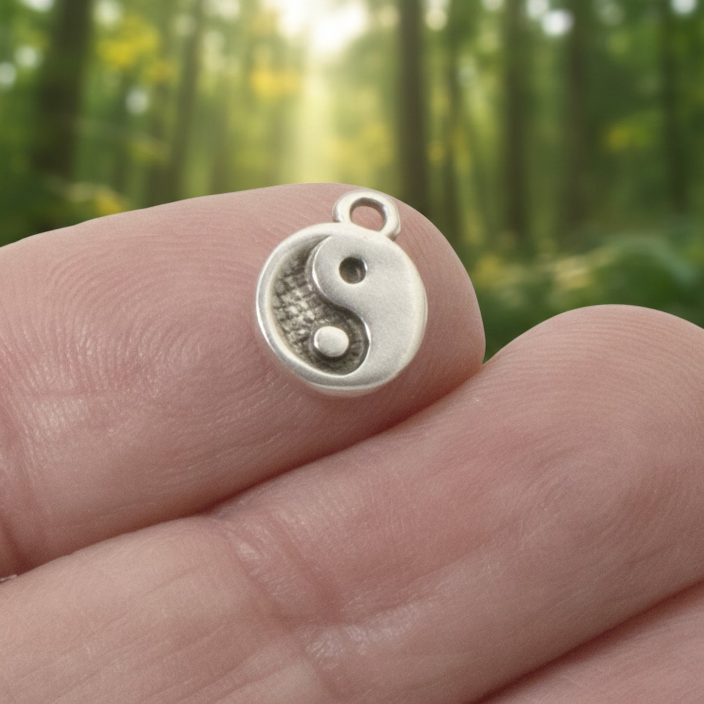 Hackberry Creek mini silver yin yang charm held between fingers against a blurred forest background.