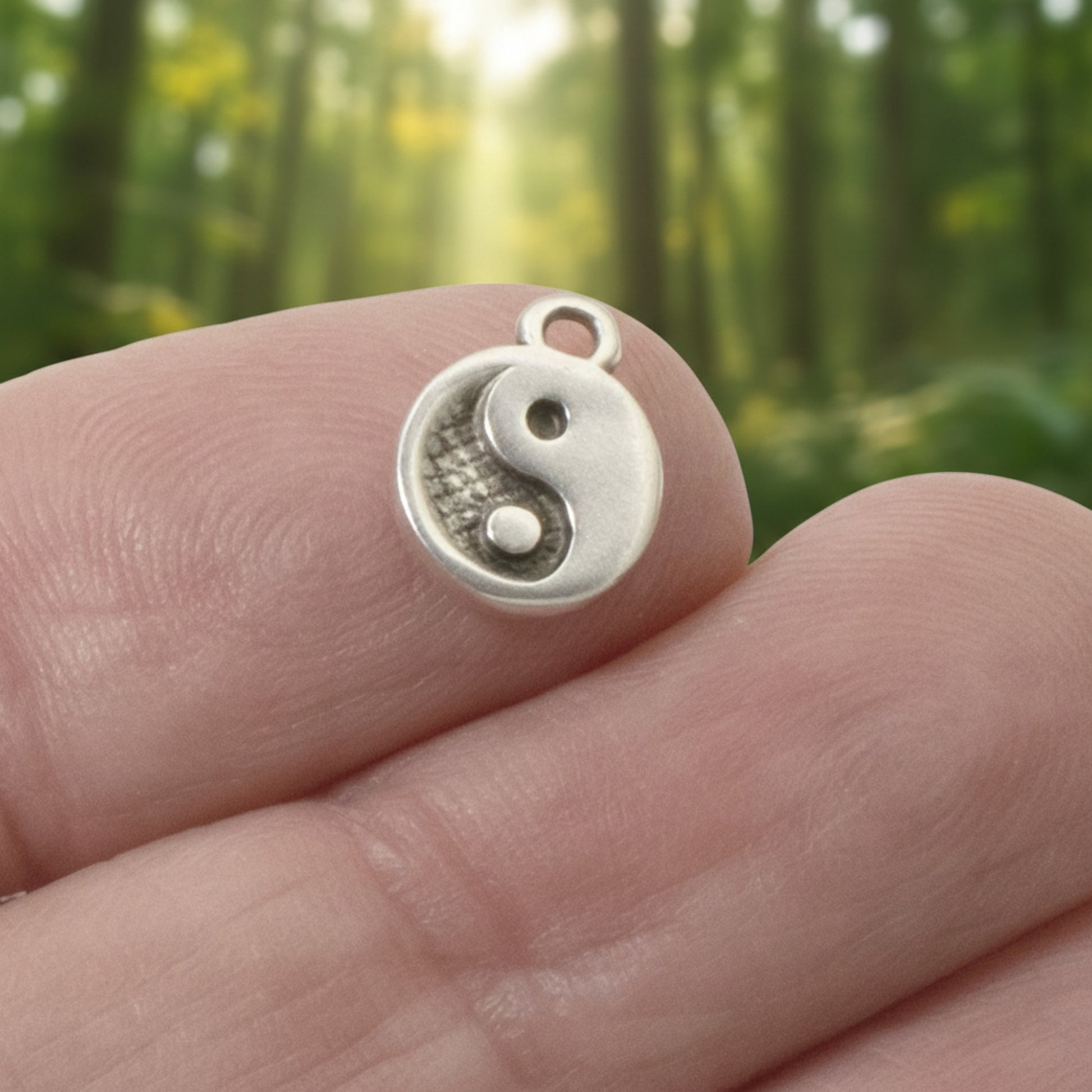 Hackberry Creek mini silver yin yang charm held between fingers against a blurred forest background.