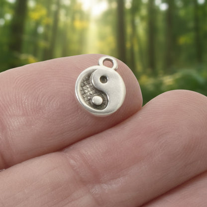 Hackberry Creek mini silver yin yang charm held between fingers against a blurred forest background.