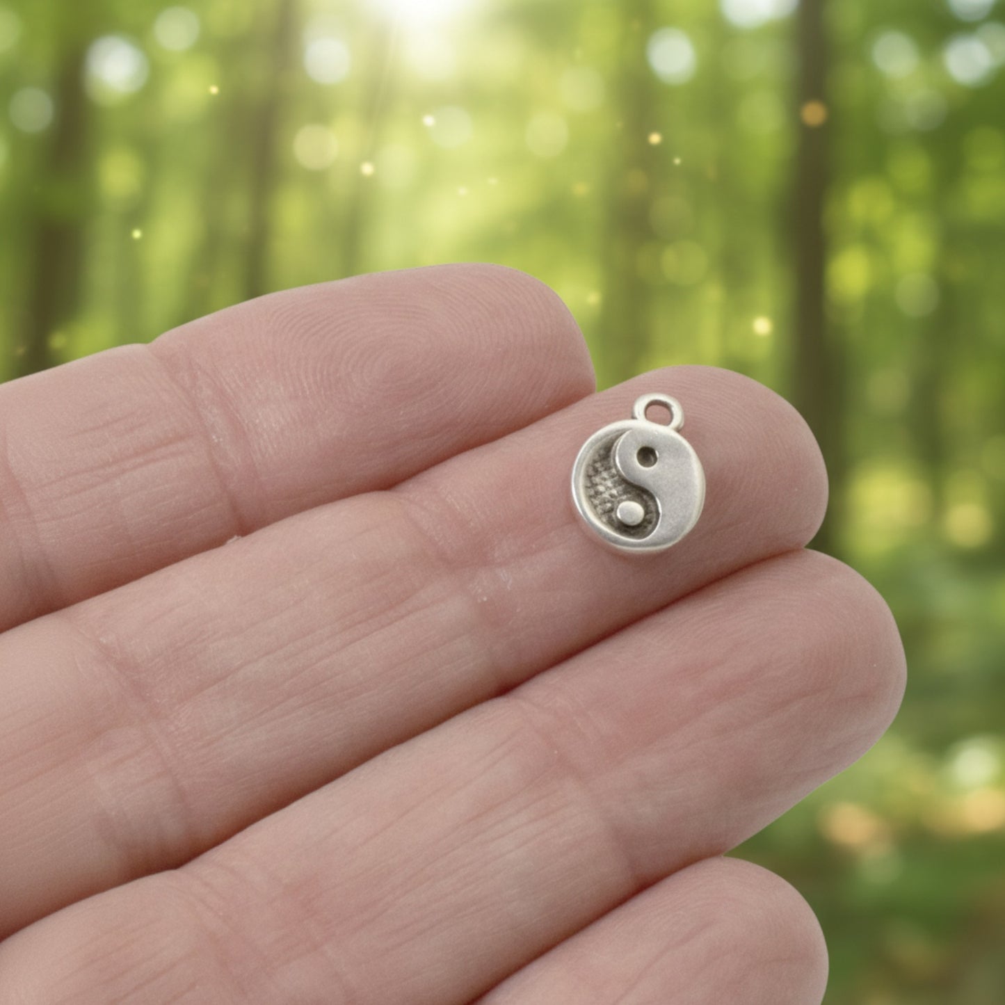 Hackberry Creek mini silver yin yang charm held on a finger against a blurred forest background