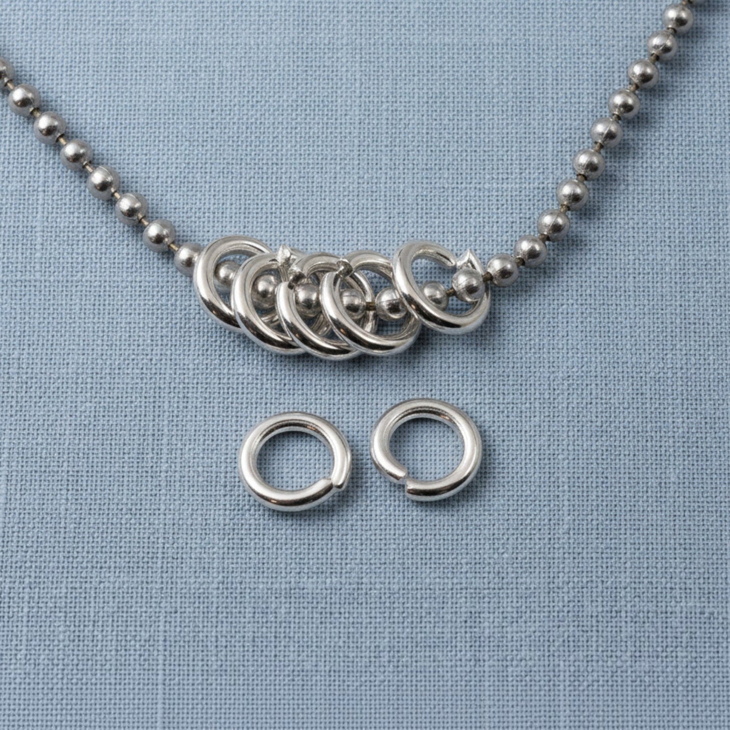 Hackberry Creek 8mm bright silver 304 stainless steel jump rings on a blue fabric background with a ball chain.