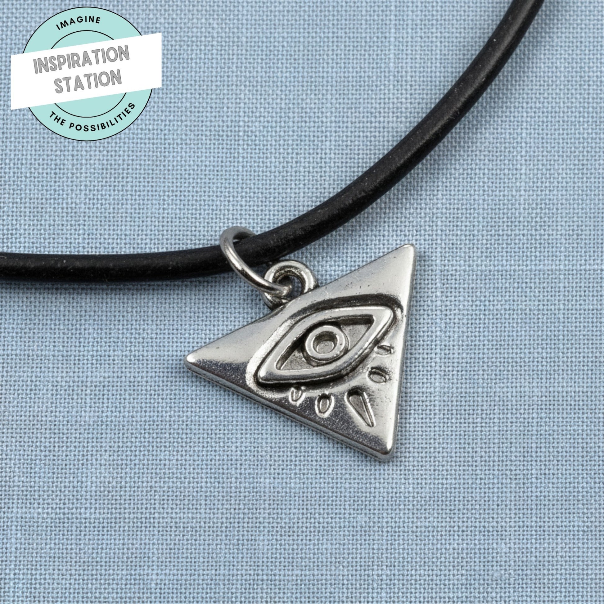 Hackberry Creek antique silver triangle evil eye charm on a black cord necklace against a blue fabric background.