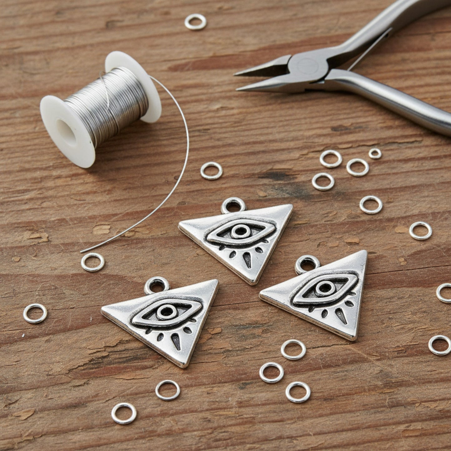 Hackberry Creek antique silver triangle evil eye charms with jump rings and wire on a rustic wooden surface.